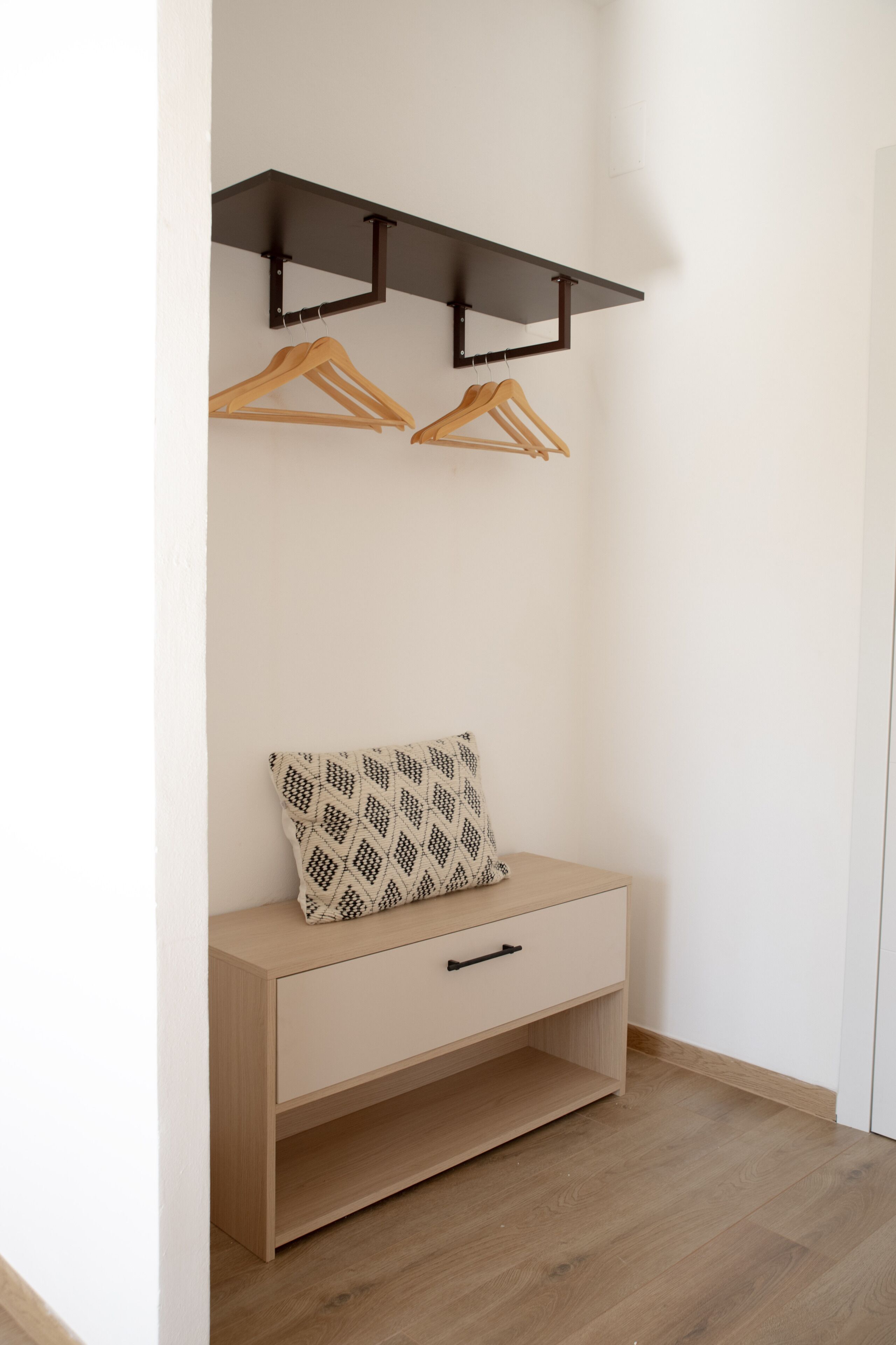 A small, neatly organized room with a wooden bench, a patterned pillow, and two wooden hangers on a black shelf.