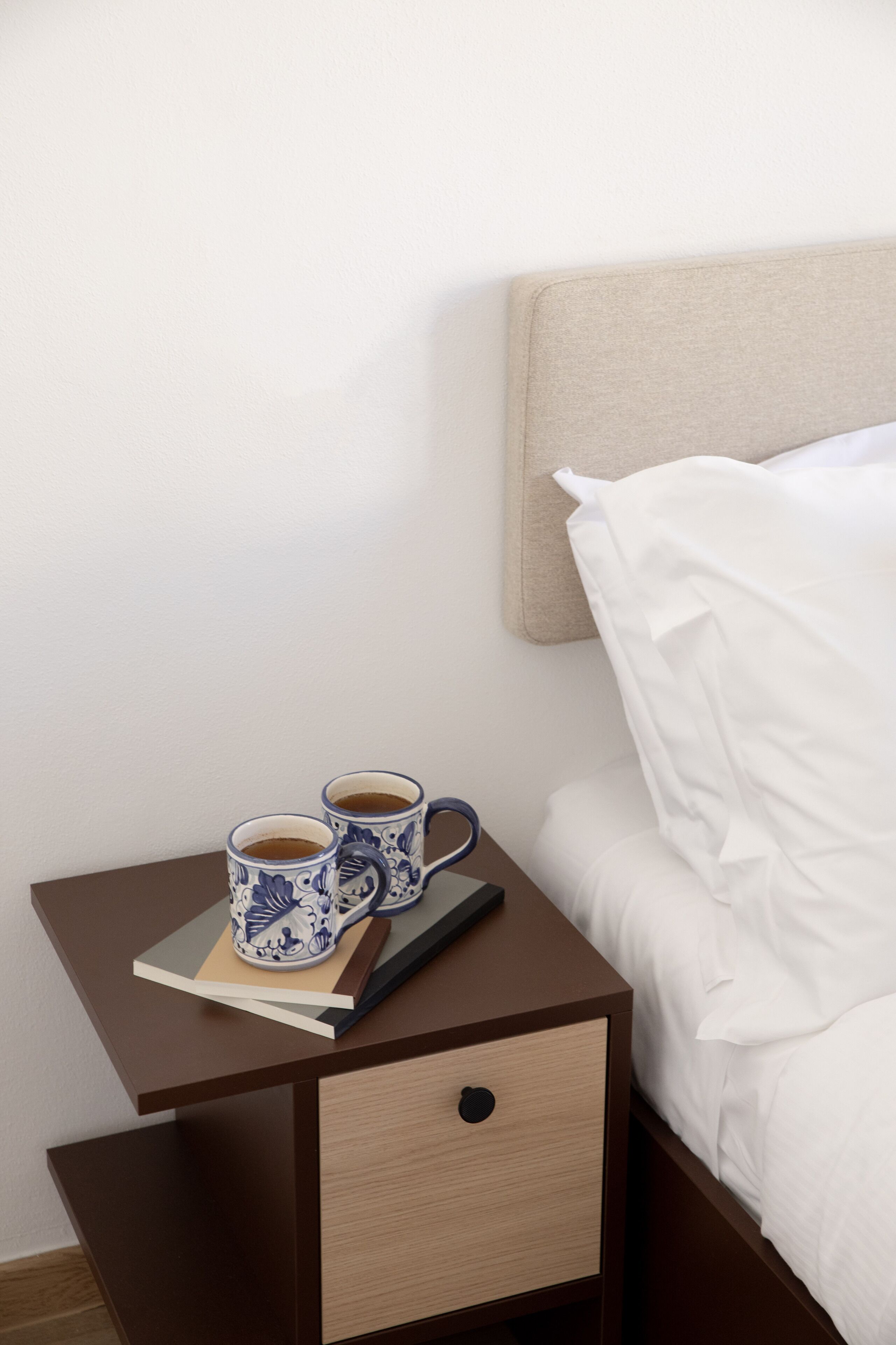 A neatly made bed with white linens, a small bedside table with a book and two cups of coffee, and a headboard against a plain wall.