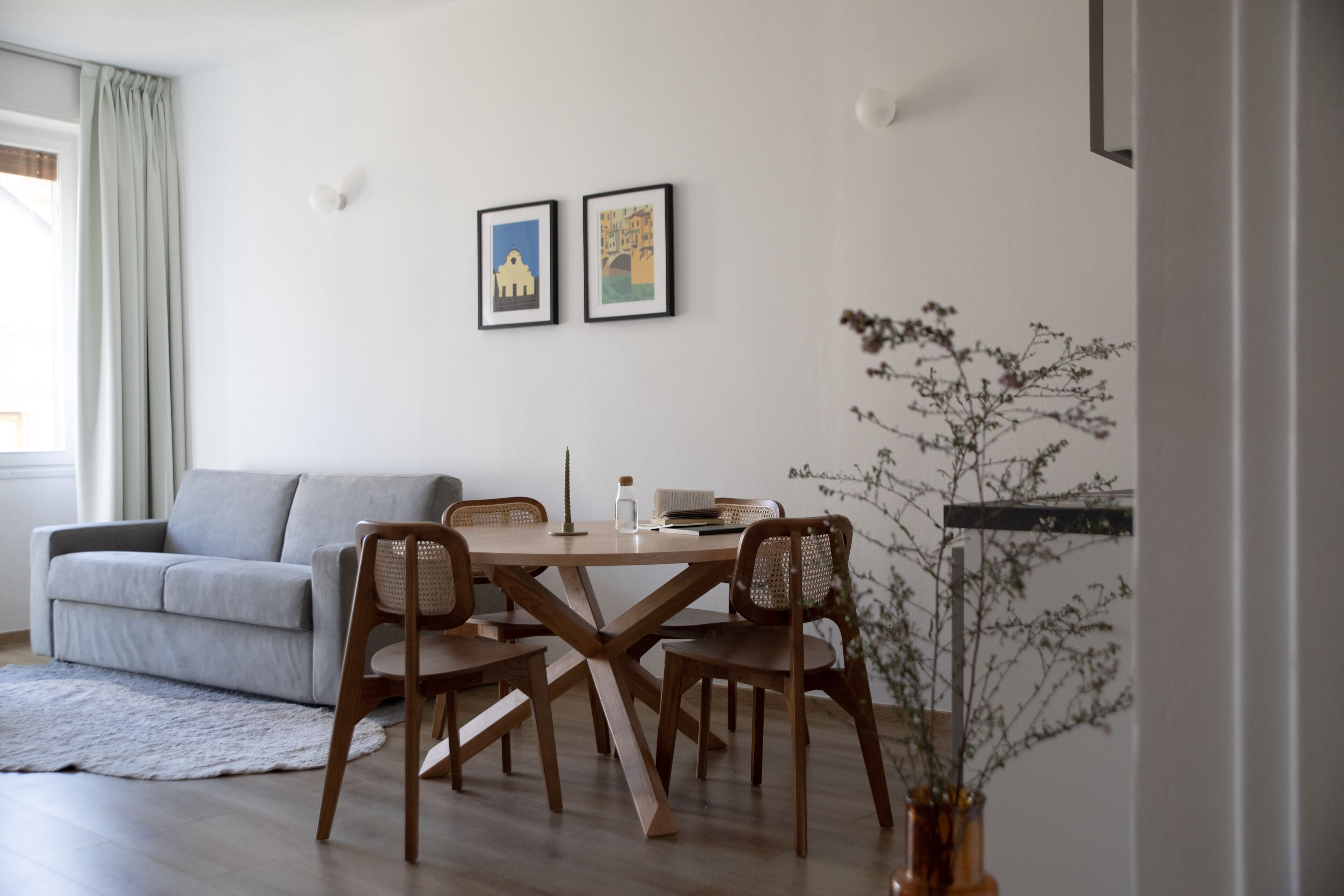 Un salon moderne avec un canapé gris, une table à manger en bois et des chaises. On peut voir deux tableaux encadrés au mur, un vase avec des branches et une fenêtre avec des rideaux.
