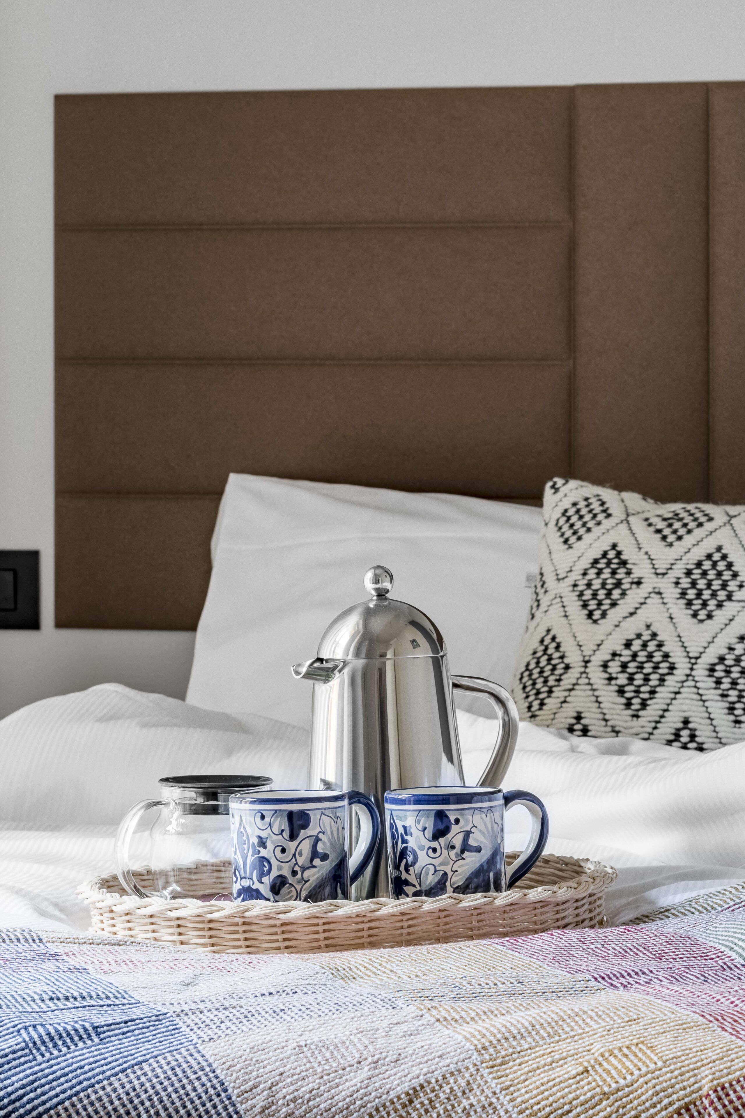 Una cama bien hecha con cabecera marrón, dos tazas estampadas, una jarra de vidrio y una bandeja tejida.