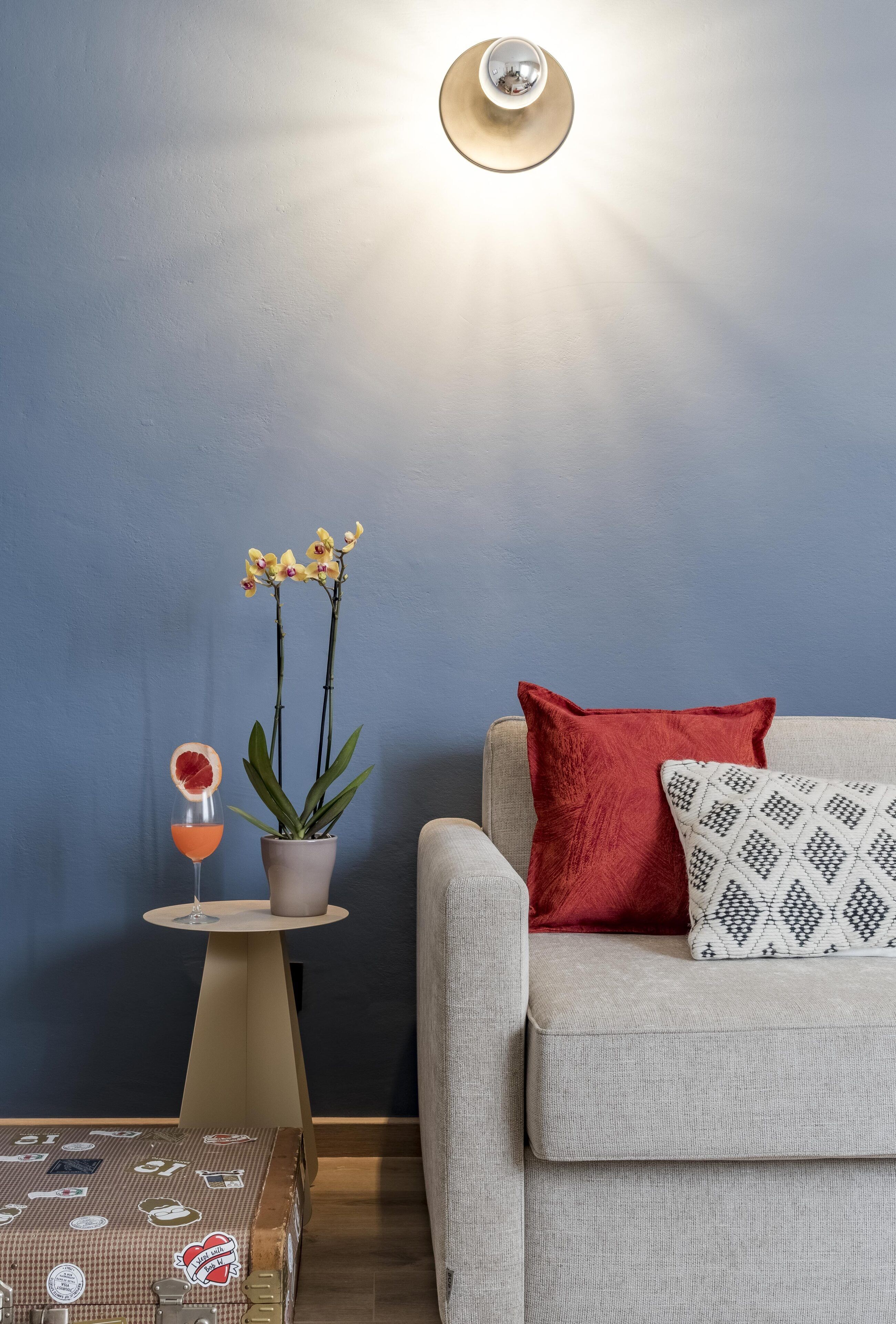 A modern living room with a grey sofa, a red pillow, a patterned pillow, a small table with a potted plant, and a glass with a fruit garnish.