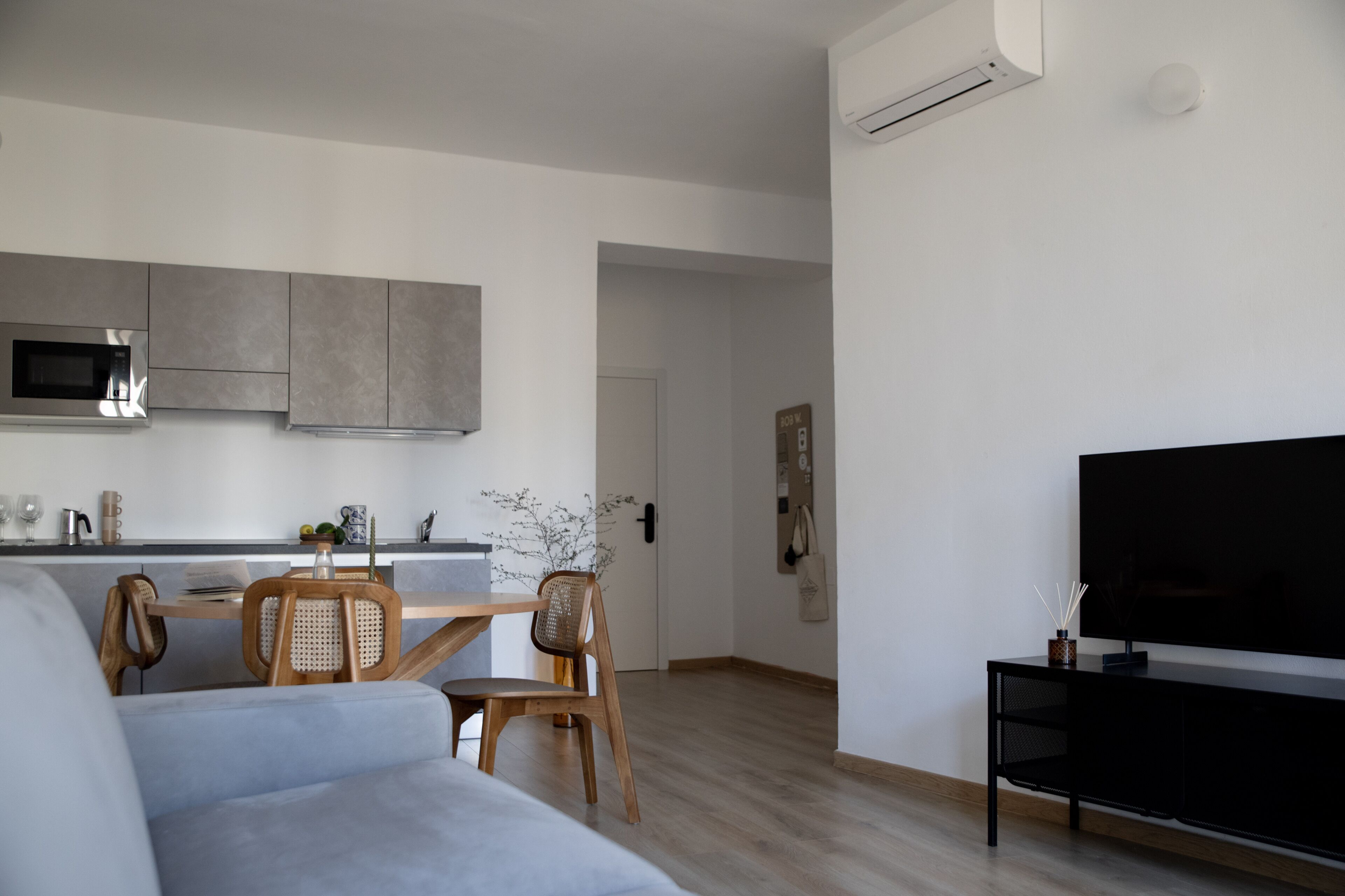 A modern living room with a grey sofa, wooden dining table with chairs, a flat-screen TV, and a kitchen area with grey cabinets and a microwave.
