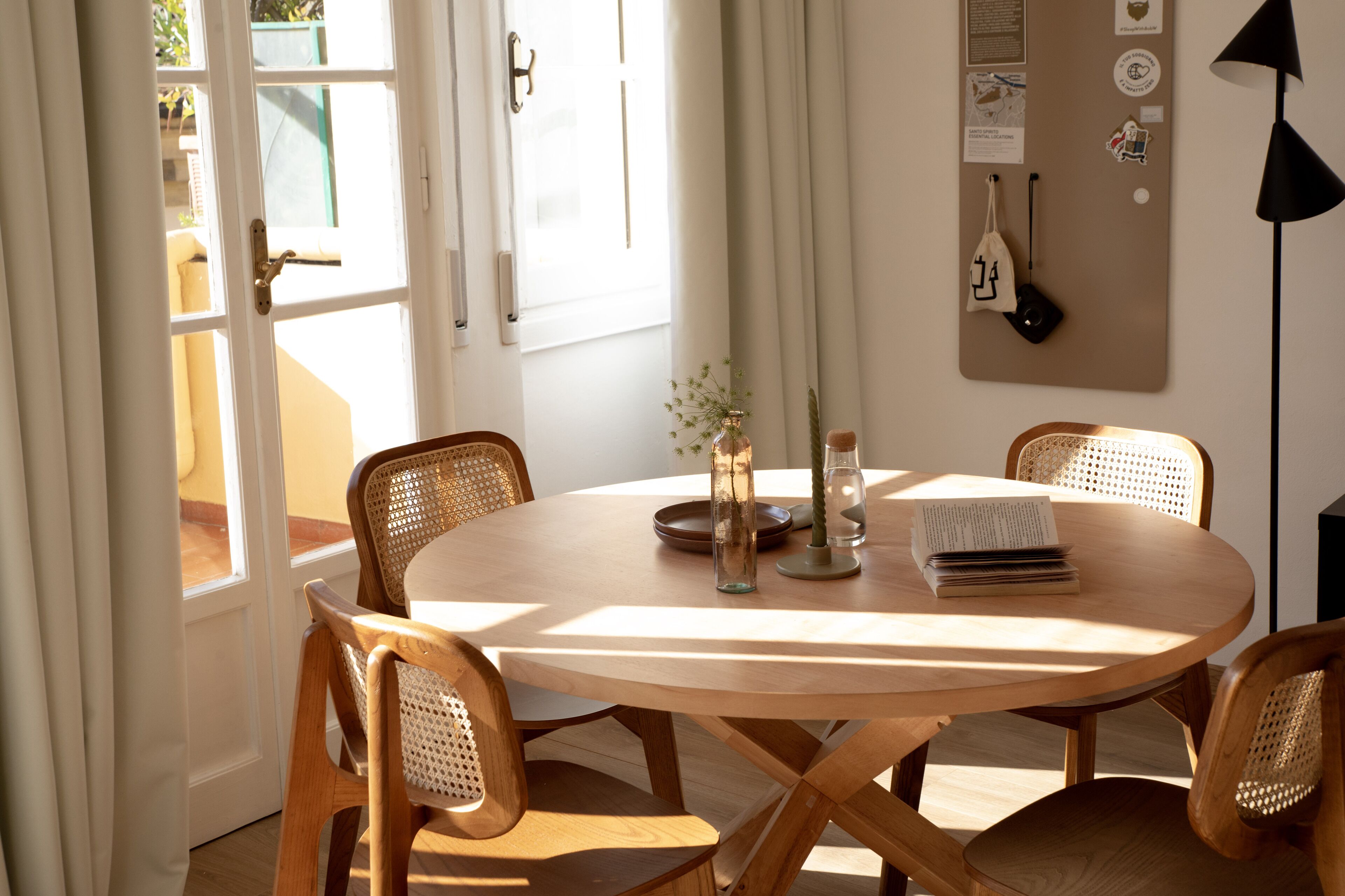 Une table ronde en bois, entourée de quatre chaises en osier, un vase en verre avec une plante, un livre et une lampe noire, le tout placé sur le côté droit.
