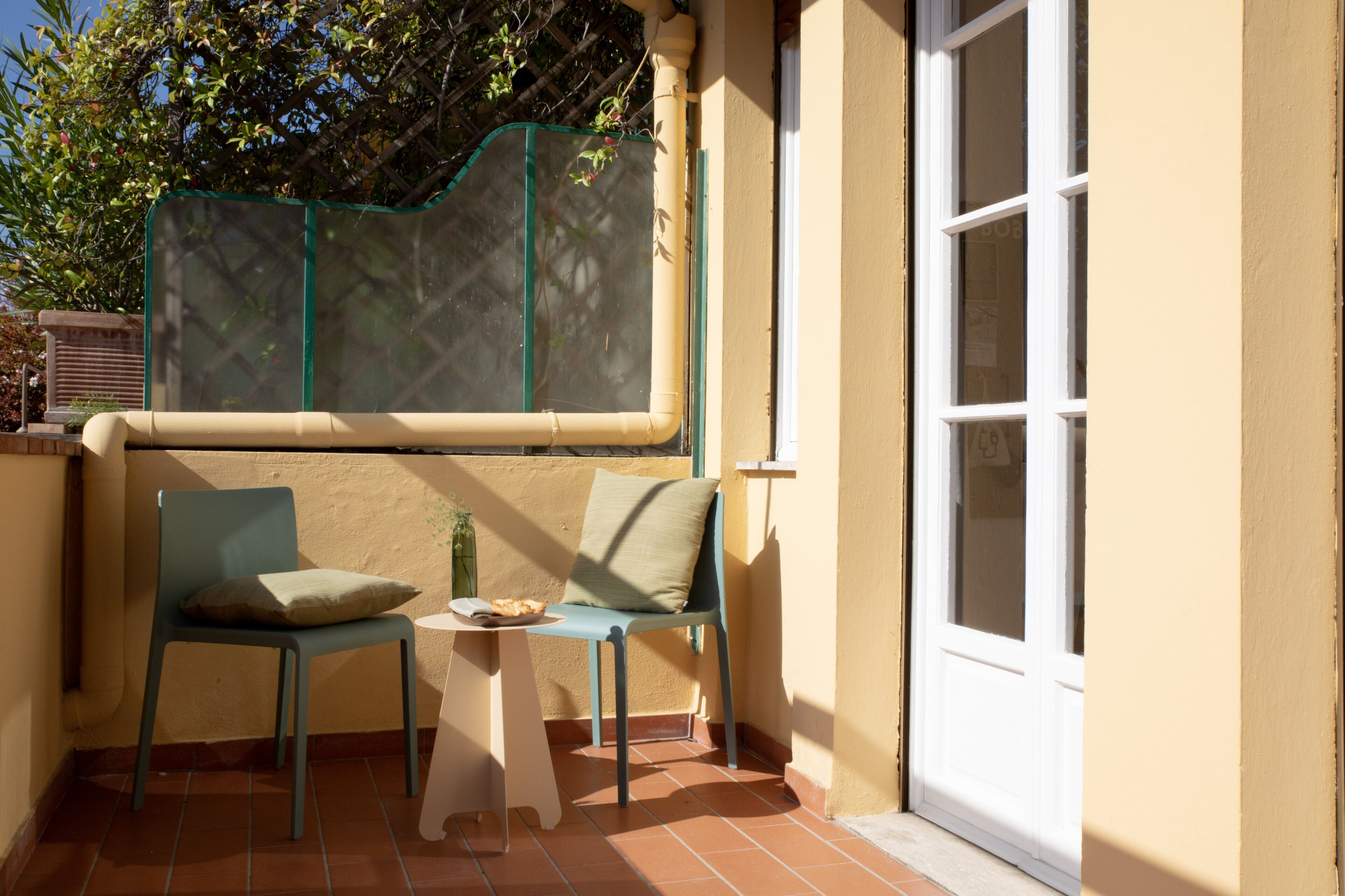 A small outdoor seating area with two chairs and a table, a window with a view of greenery, and a door to the right.