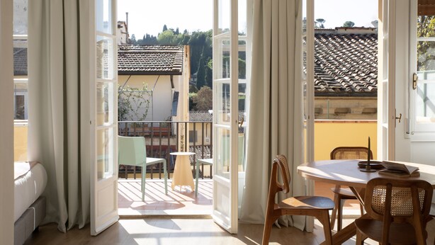 Een open balkon met een tafel en stoelen, een uitzicht op een pannendak, en een deur die naar de binnenkant leidt.