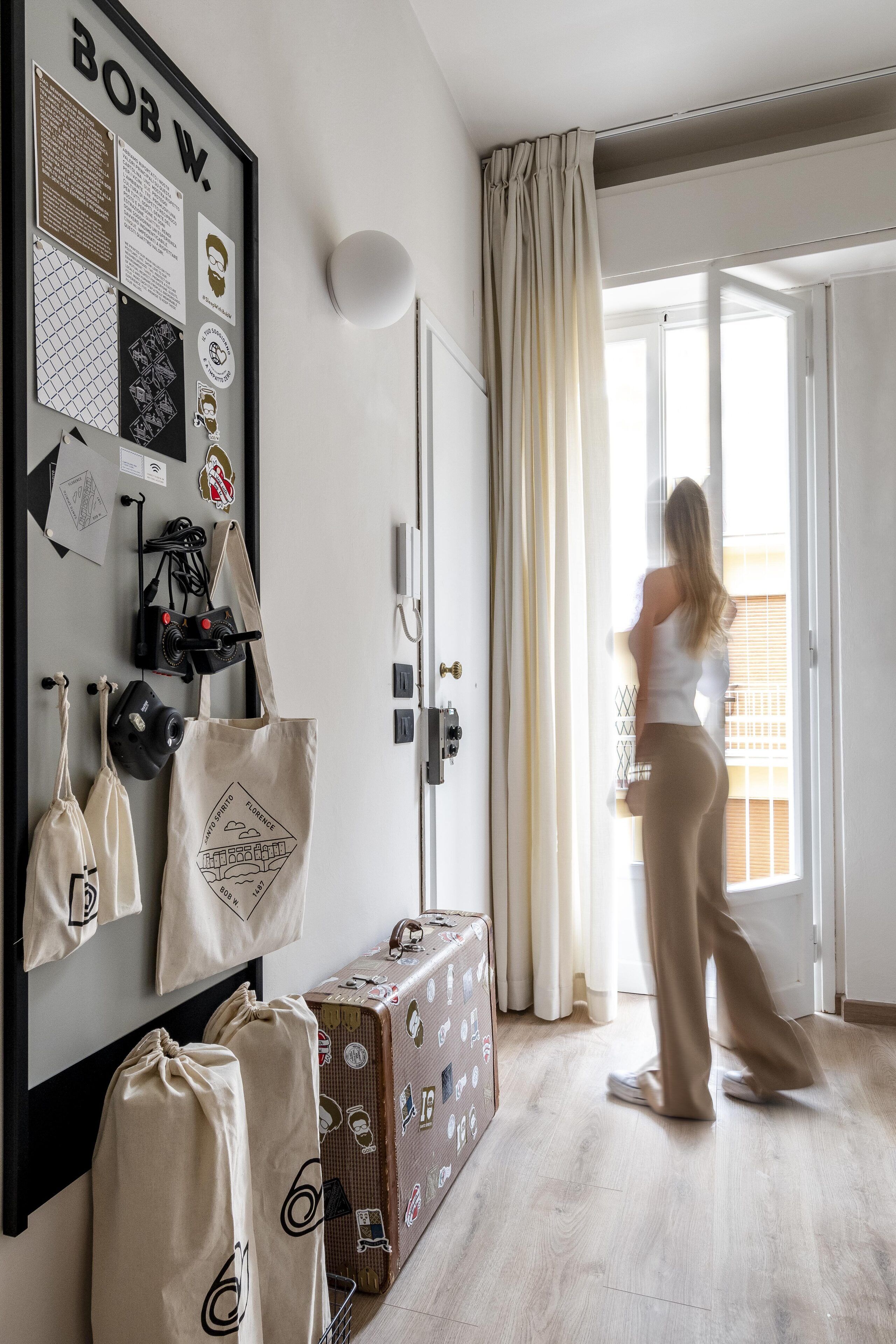 Une pièce avec un grand miroir, une armoire, une valise et une personne debout près d’une fenêtre.