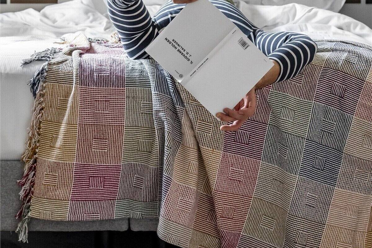 A person lying on a bed with a patterned blanket, holding a book titled 'The Little House on the Prairie.'
