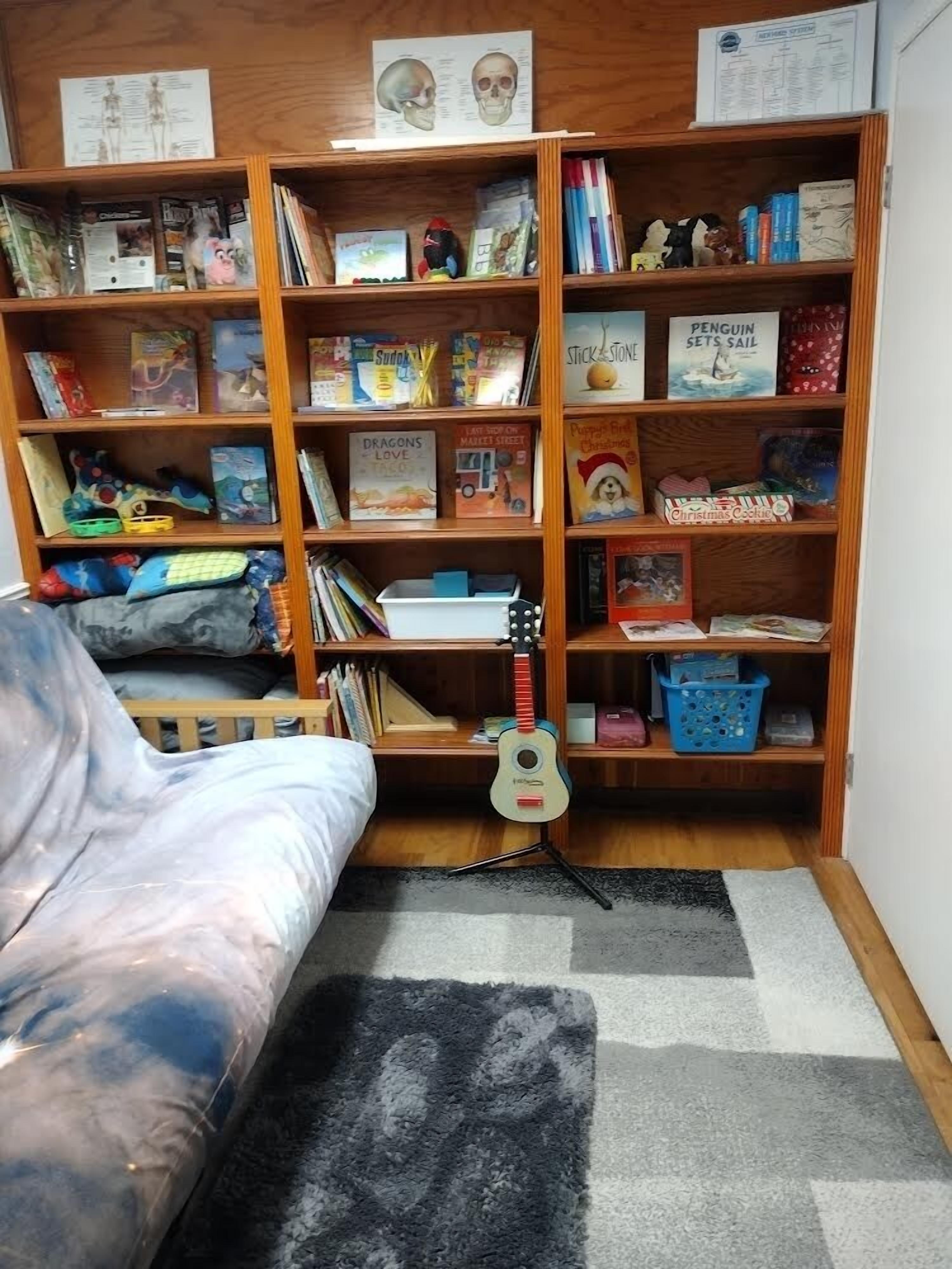 Et hyggeligt værelse med en træreol fyldt med bøger, en guitar og diverse legetøj.
