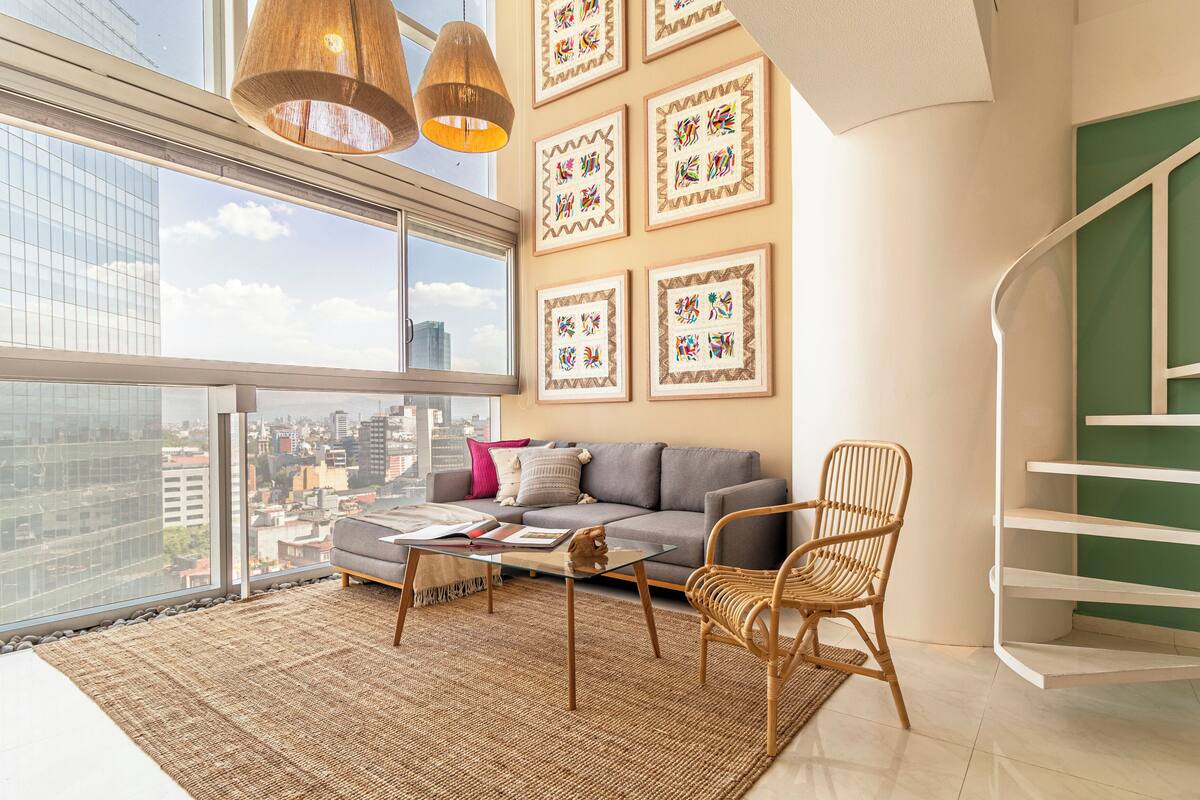 A modern living room with a city view, a gray sofa, a wicker chair, a glass coffee table, and a staircase with a green accent wall.