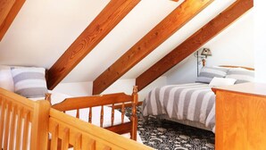 Un dormitorio con techo inclinado, vigas de madera, cama con ropa de cama a rayas, una mesita de noche y una lámpara.