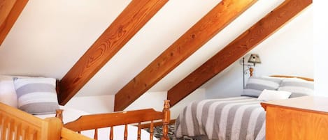 A bedroom with a sloped ceiling, wooden beams, a bed with striped bedding, a nightstand, and a lamp.