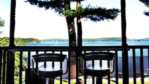 Una terraza de madera con dos sillas de metal y una mesa, con vistas a un lago rodeado de árboles y un cielo despejado.