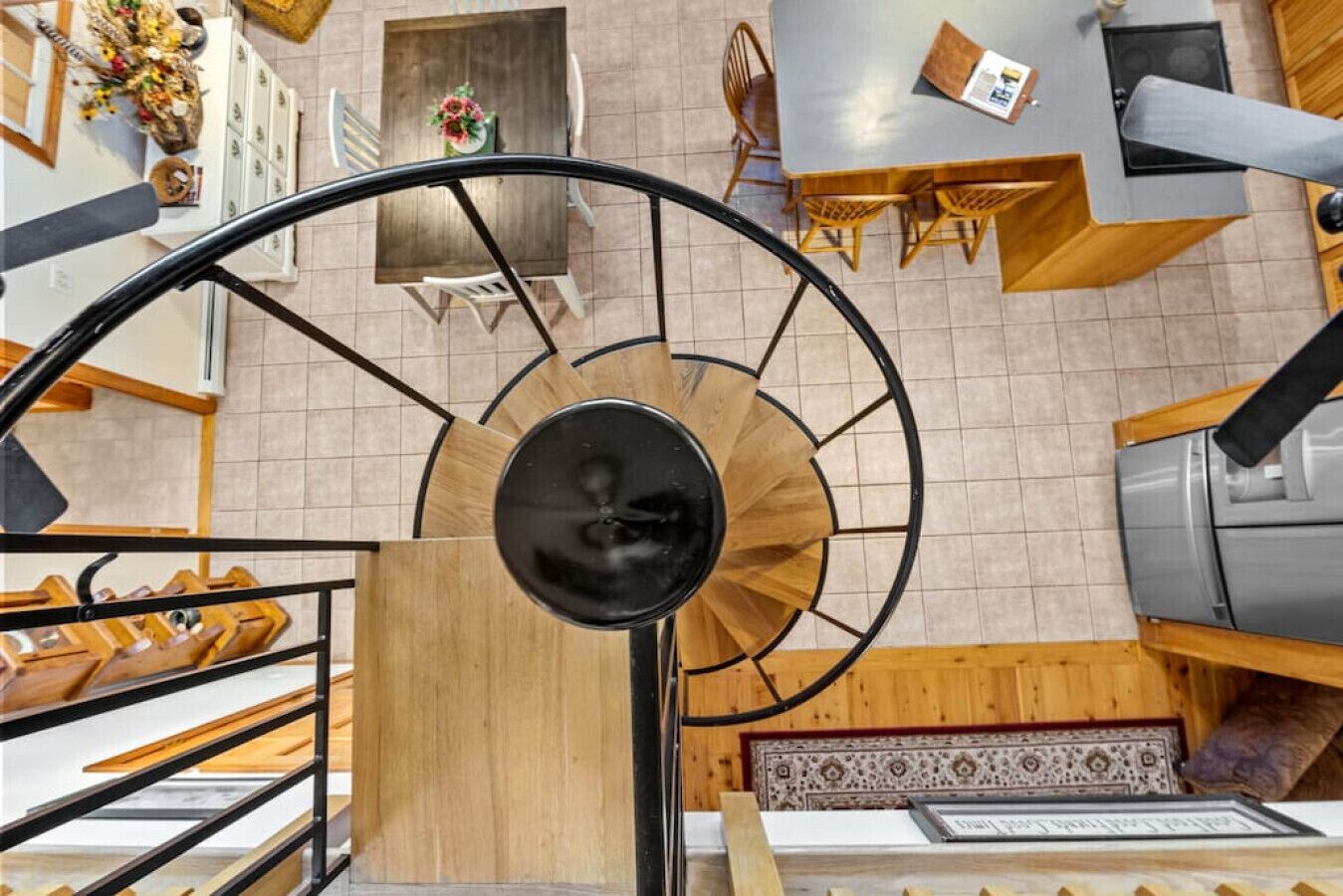 A spiral staircase with a wooden finish and black metal railings, leading to a dining area with wooden tables and chairs.