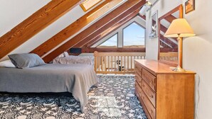 A bedroom with a sloped ceiling, wooden beams, a bed with pillows, a dresser, a lamp, and a patterned rug. - Cottage · Beachfront Cottage on Lake Champlain, Colchester (Colchester)