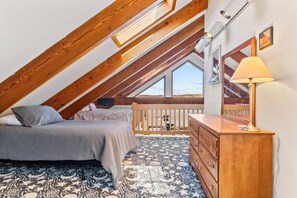 A bedroom with a sloped ceiling, wooden beams, a bed with pillows, a dresser, a lamp, and a patterned rug. - Cottage · Beachfront Cottage on Lake Champlain, Colchester (Colchester)