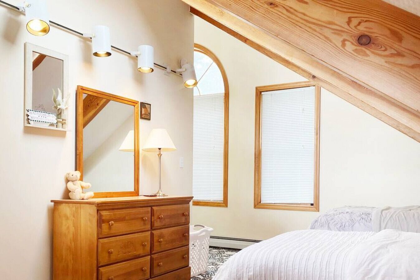 A bedroom with a wooden dresser, a mirror, a lamp, and a bed. There are two windows with blinds and a sloped ceiling.