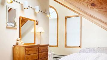 A bedroom with a wooden dresser, a mirror, a lamp, and a bed. There are two windows with blinds and a sloped ceiling.