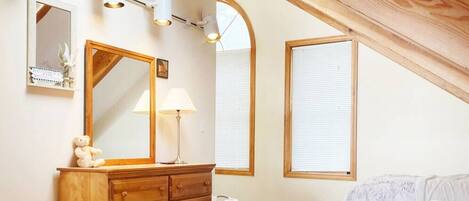 Une chambre à coucher avec une commode en bois, un miroir, une lampe et un lit. Il y a deux fenêtres équipées de stores et un plafond en pente.