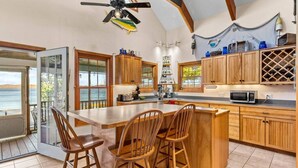 Una cocina con armarios de madera, una isla central con taburetes de bar, un ventilador de techo y vistas al mar a través de amplias ventanas.