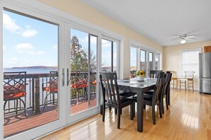 Un comedor amplio con una gran mesa y sillas, vistas al océano a través de puertas corredizas de vidrio, y una zona de cocina con refrigerador.
