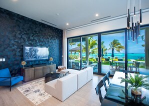 A modern living room with a large TV, a sofa, and a dining area with a view of the ocean. - Luxury 4 Bedroom in Bodden Town (Breakers)