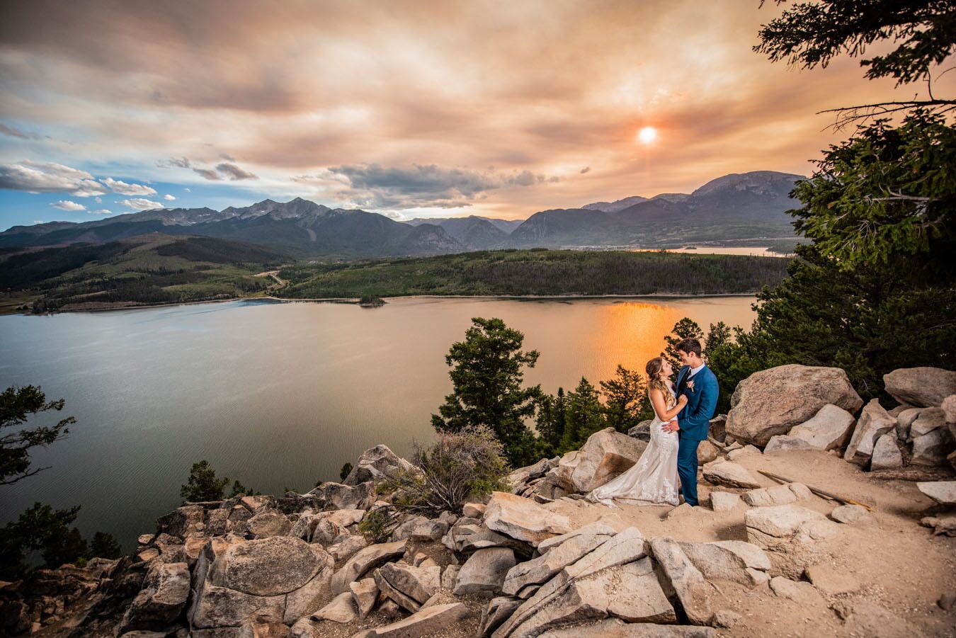 A couple embracing on a rocky overlook with a lake and mountains in the background.