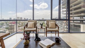 A high-rise room with a large window, two chairs, a table, a book, and a vase with flowers. - Miyana by Kukun (Mexico City)
