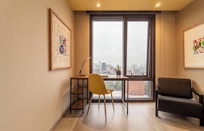 Une pièce comprenant un bureau, une chaise jaune, un canapé foncé, une grande fenêtre donnant sur la ville et des œuvres d’art encadrées accrochées aux murs.