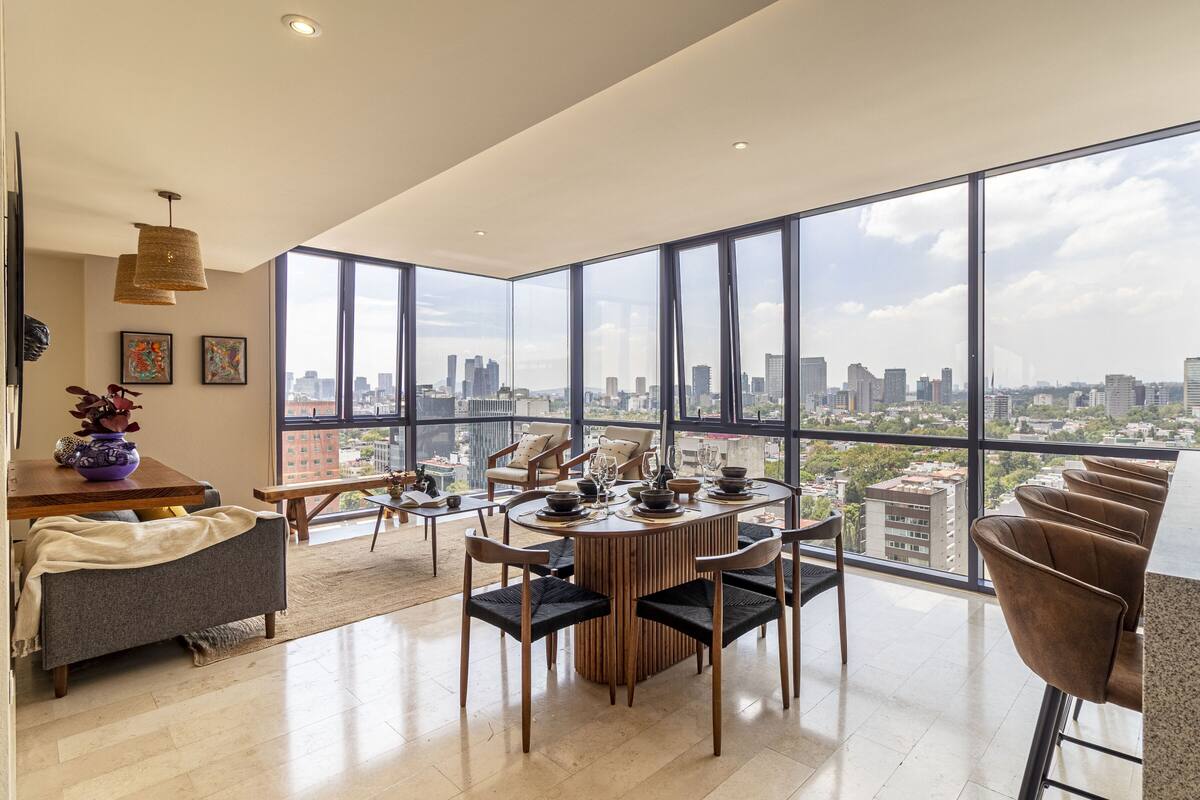 A modern dining area with a panoramic city view, featuring a round table with chairs, a coffee table, and a vase with flowers.