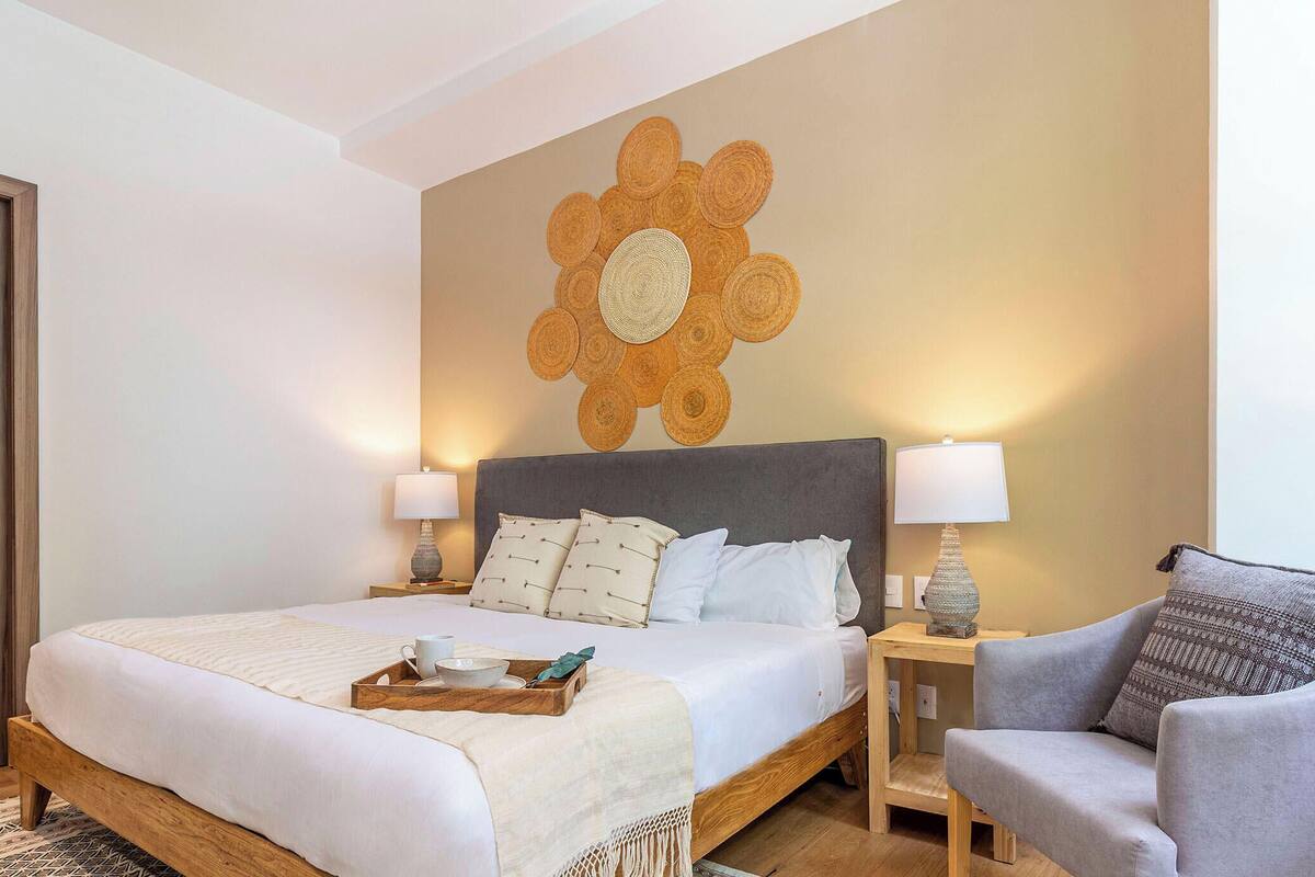 Une chambre d’hôtel avec un lit, deux tables de chevet avec des lampes, un fauteuil gris, un plancher en bois et un grand élément décoratif mural.
