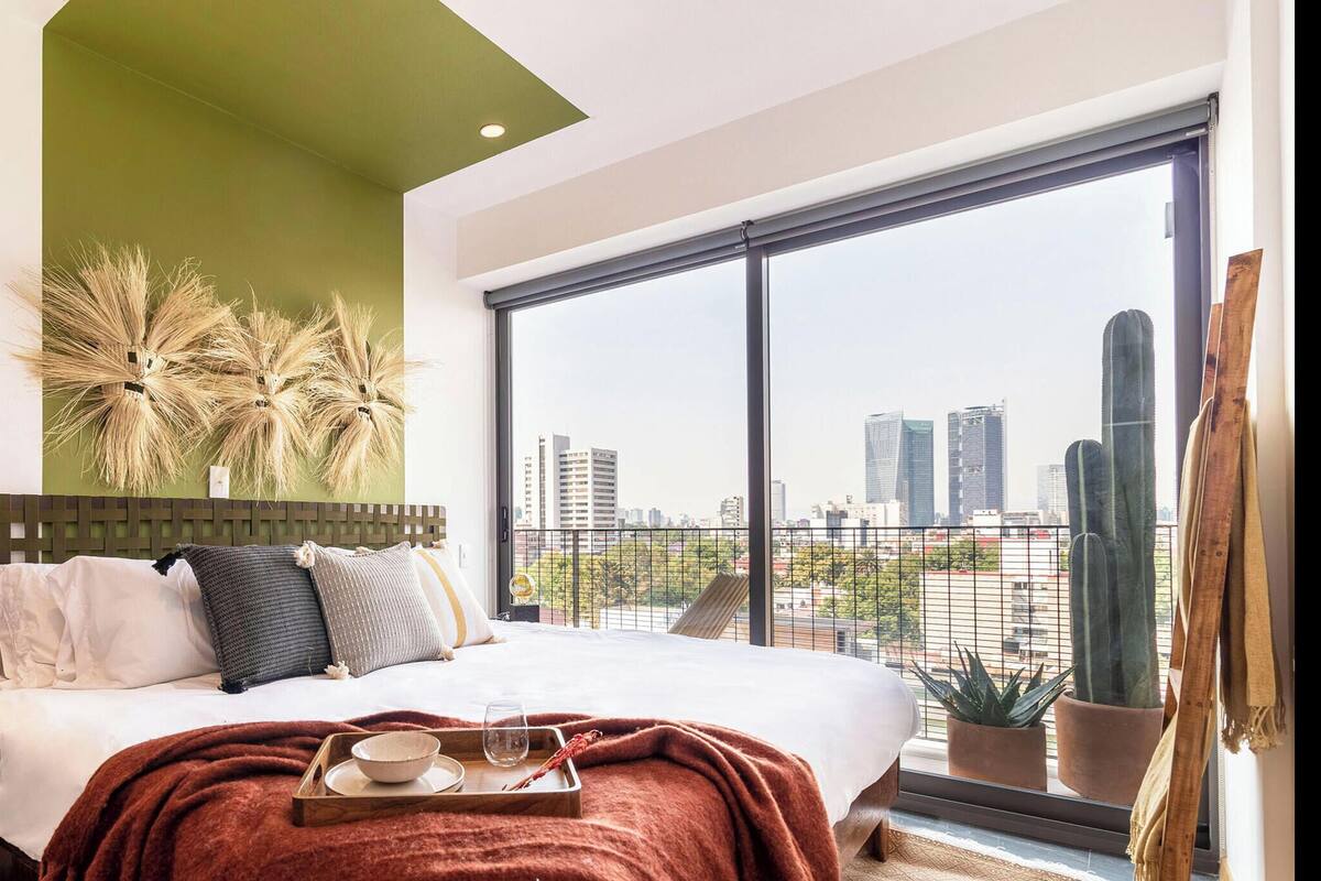 Une chambre avec un grand lit, une vue sur la ville, un mur d’accent de couleur verte et un cactus.