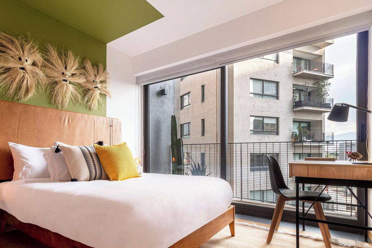 Une chambre moderne avec un grand lit, une table de chevet avec une lampe et une vue sur la ville depuis un balcon.