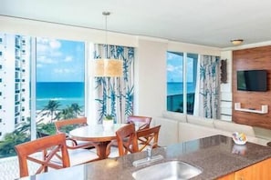 Una cocina moderna con un ventanal grande con vista al océano, una zona de comedor con sillas de madera y un televisor empotrado.