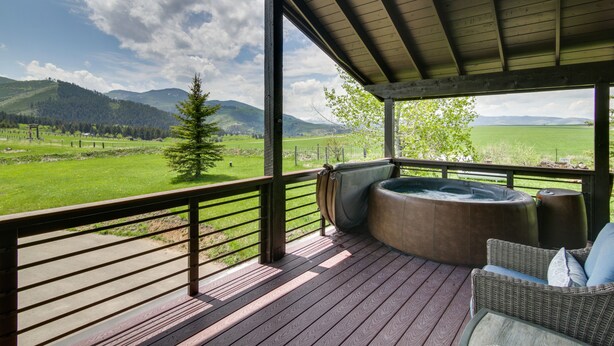 A covered outdoor deck with a hot tub and wicker furniture, overlooking a green landscape and mountains. - Stunning Gallatin Gateway Mountain Retreat! (Gallatin Gateway)