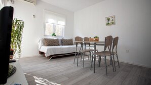 A modern living room with a sofa, dining table, and chairs. There is a TV on the left, a window with blinds, and a plant.