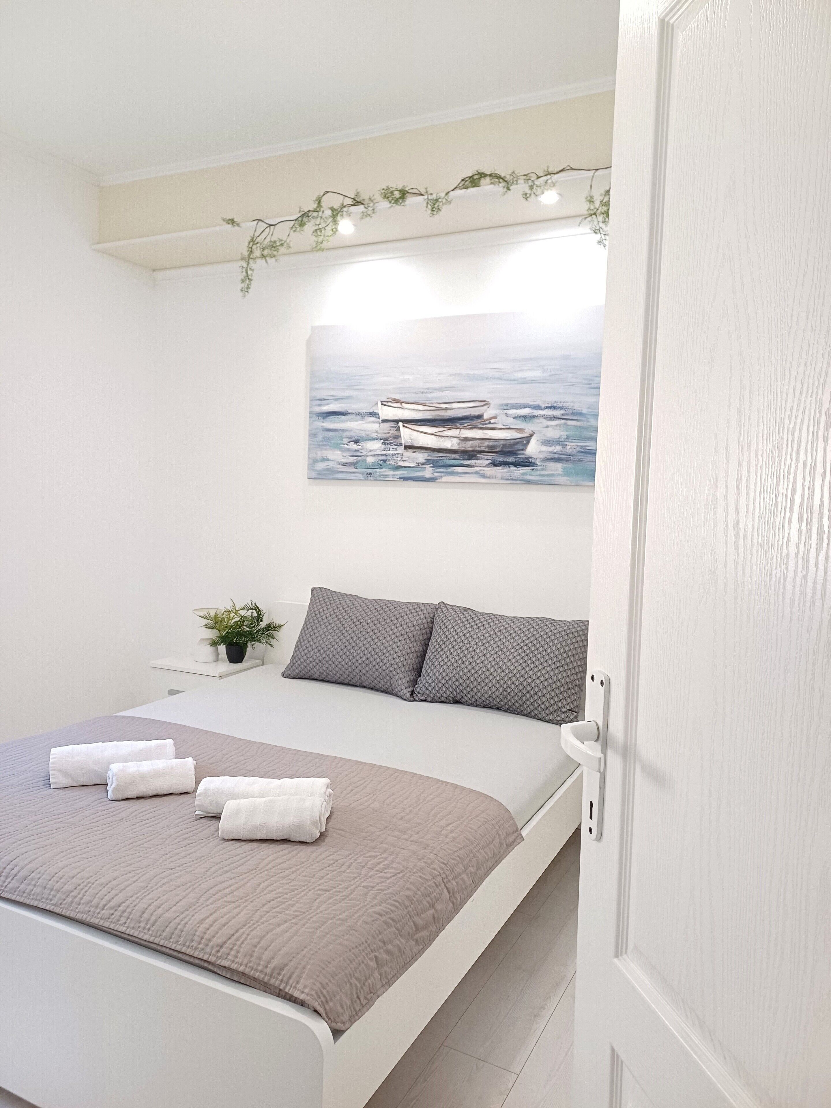 Un dormitorio con cama, almohadas, un cuadro de barcos, una planta pequeña y una puerta.