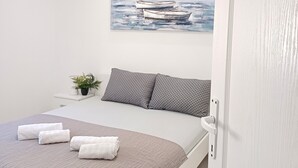 A bedroom with a bed, pillows, a painting of boats, a small plant, and a door.