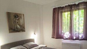A bedroom with a bed, a nightstand, a wall-mounted heater, a window with curtains, and a painting on the wall.