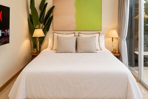 Un lit bien fait, recouvert d’une couvre-lit blanche et agrémenté de deux oreillers, est encadré par deux tables de chevet sur lesquelles reposent des lampes. Au-dessus du lit, on peut admirer une grande peinture abstraite verte, et une fenêtre avec des rideaux se trouve à droite.