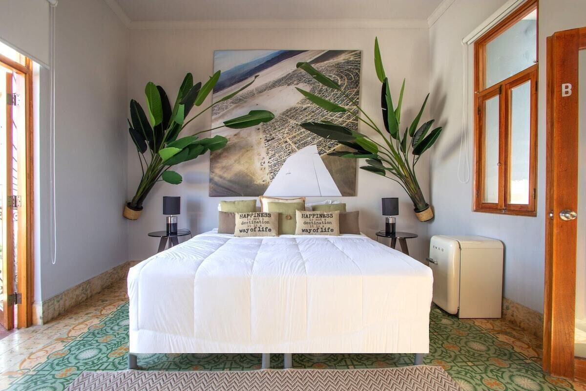 A bedroom with a large bed, two potted plants, a painting, and a small refrigerator.