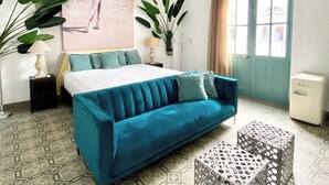 A bedroom with a bed, a blue sofa, a patterned coffee table, a ceiling fan, a large window, and a painting on the wall.