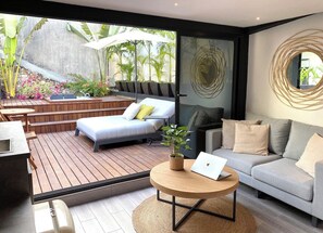 A modern living room with a sofa, a round wooden coffee table with a laptop, a bed, and a view of a garden with plants and a concrete wall.