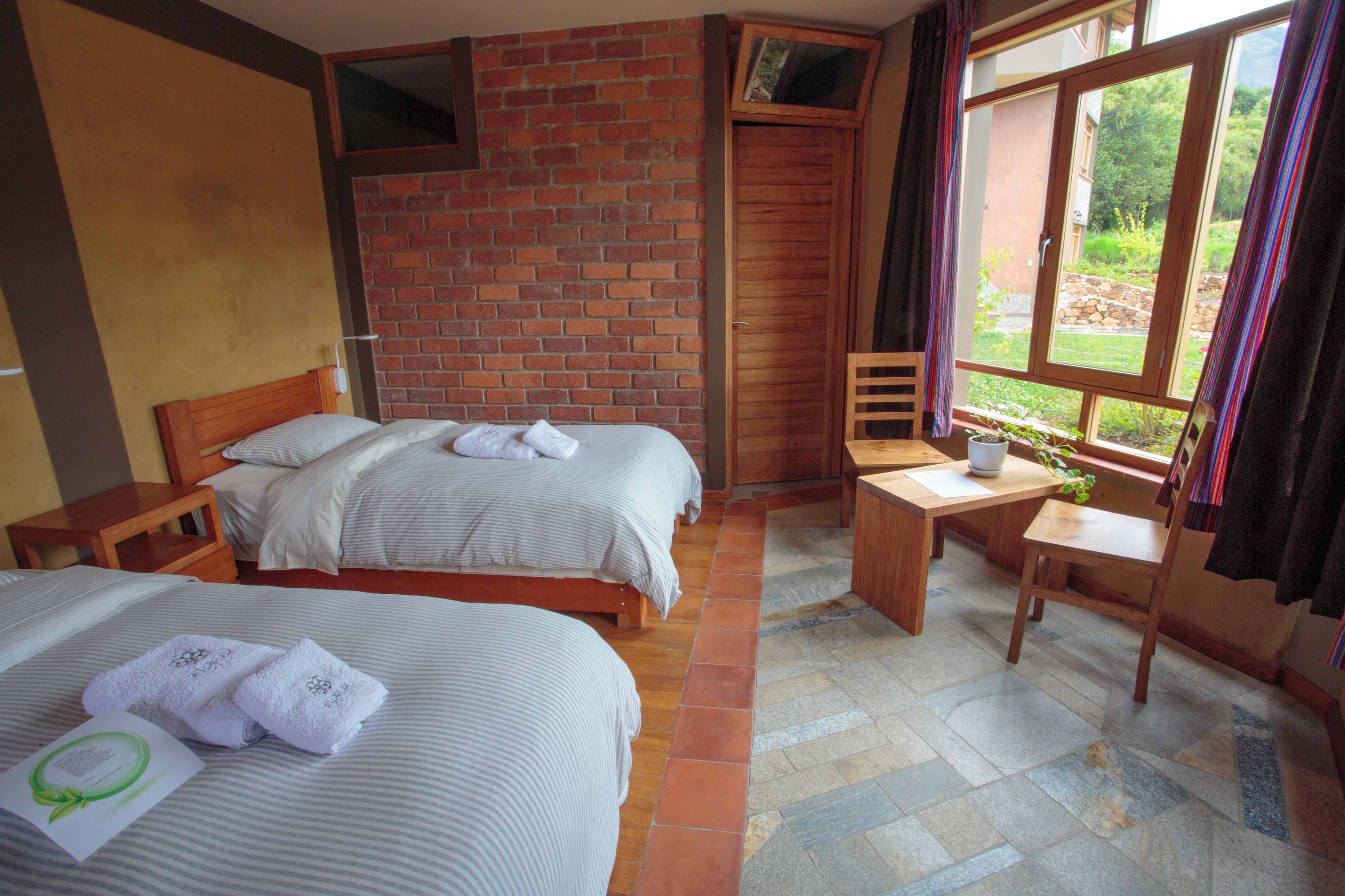 A hotel room with two beds, a wooden desk, a chair, a window with curtains, and a brick wall.