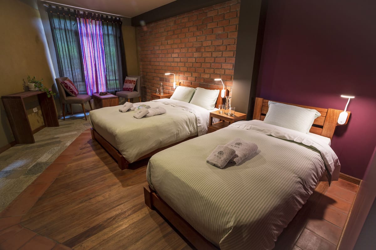 A hotel room with two beds, a brick wall, and a window with colorful curtains.