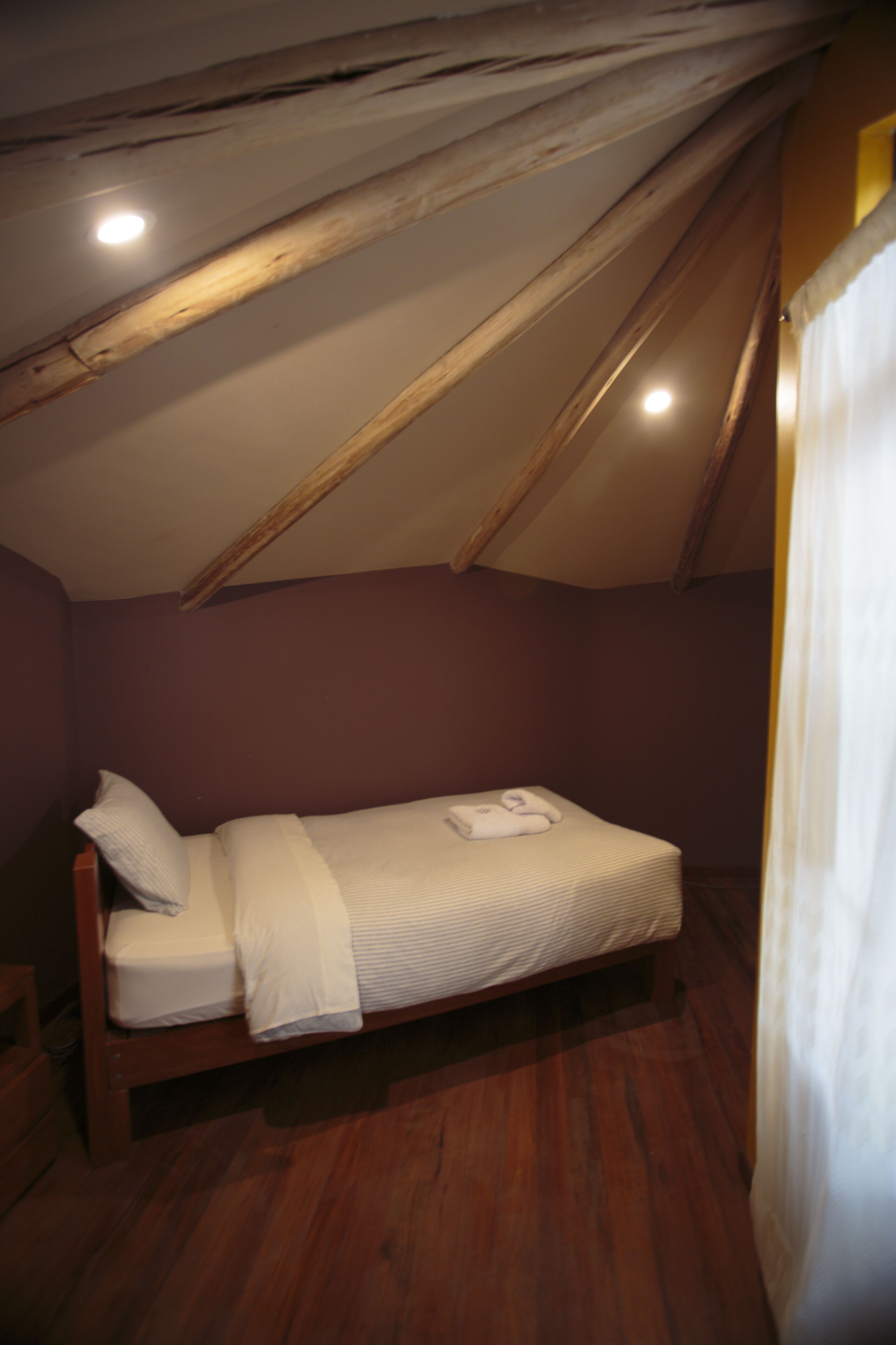 Une petite chambre mansardée, peu éclairée, avec un lit simple, des poutres en bois au plafond et un mur uni.