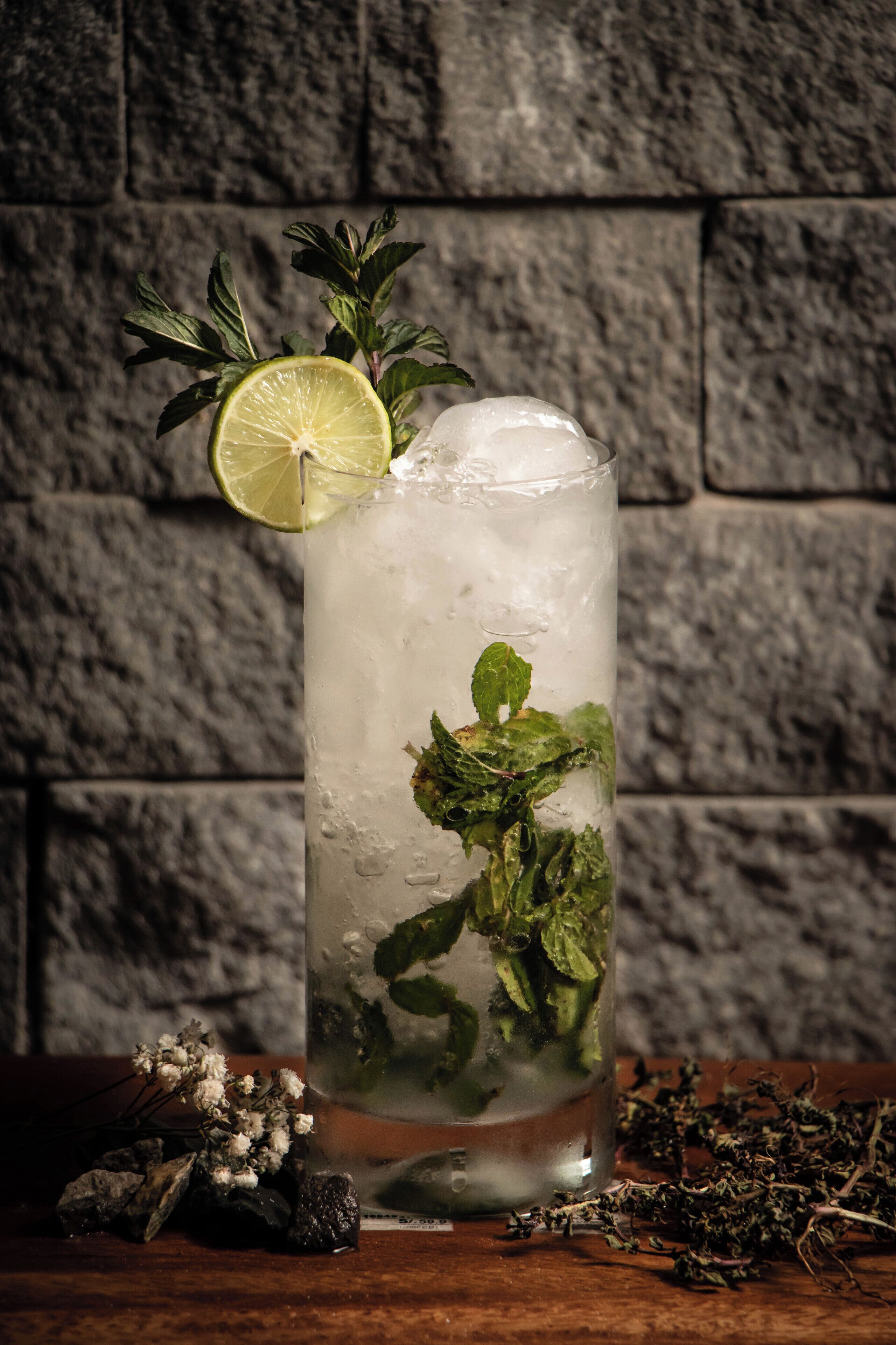 A tall glass of iced beverage with mint leaves and a lime slice, set against a stone wall background.