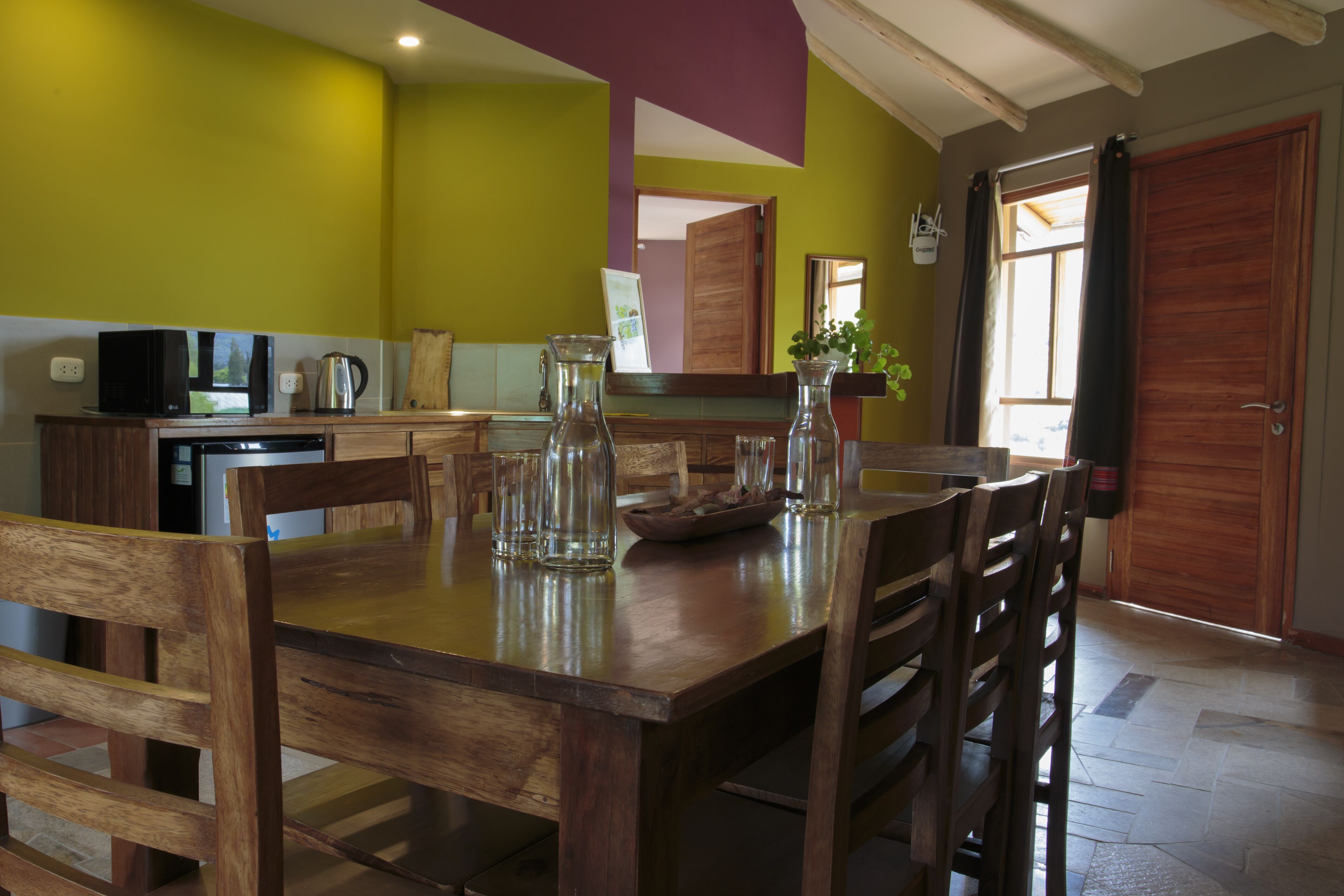Una mesa de comedor de madera con sillas, una zona de cocina con microondas, hervidor y armarios, y una puerta grande de madera.