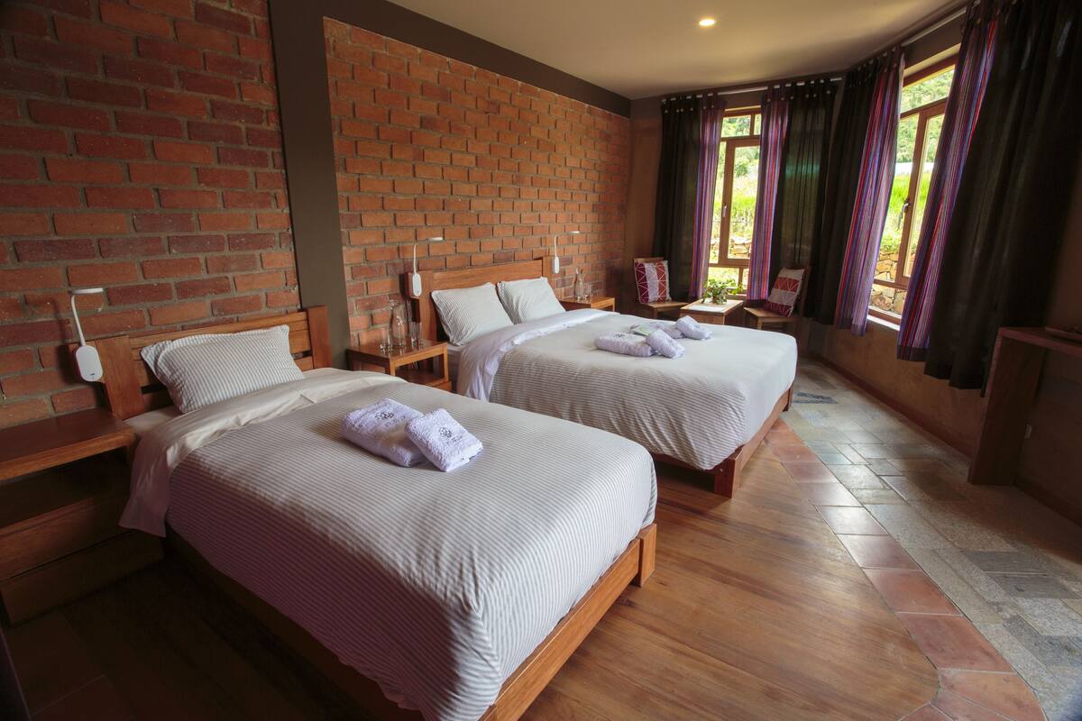 A hotel room with two beds, a brick wall, wooden flooring, and a view through large windows.