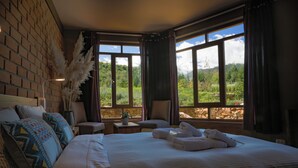 A hotel room with a large bed, two chairs, a small table with a plant, and a view of a mountainous landscape through large windows. - Fleur Lodge (Acopampa)