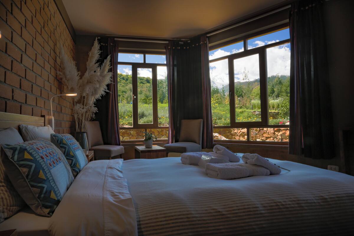 A hotel room with a large bed, two chairs, a small table with a plant, and a view of a mountainous landscape through large windows.